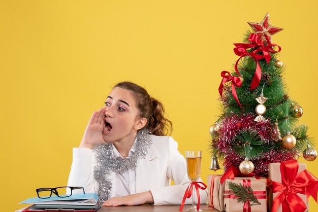 Vista frontal doctora sentada detrás de su mesa sobre fondo amarillo con árbol de navidad y cajas de regalo