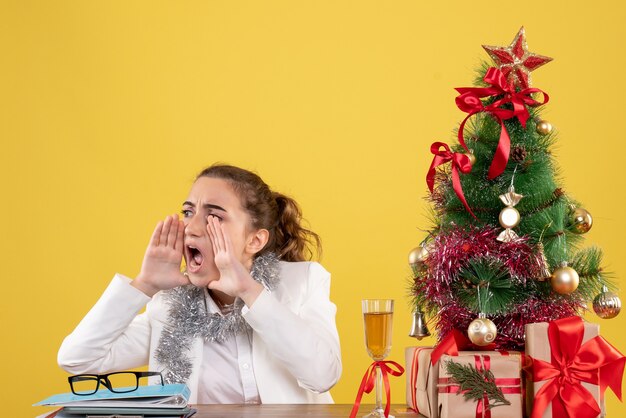 Vista frontal doctora sentada detrás de su mesa sobre fondo amarillo con árbol de navidad y cajas de regalo