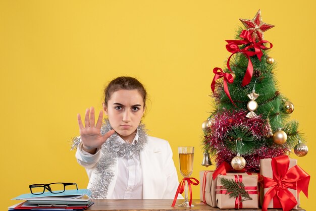 Vista frontal doctora sentada detrás de su mesa sobre fondo amarillo con árbol de navidad y cajas de regalo