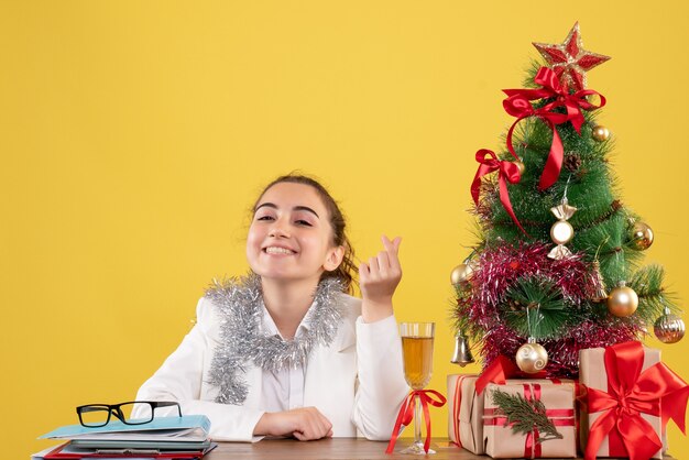 Vista frontal doctora sentada detrás de su mesa sobre fondo amarillo con árbol de navidad y cajas de regalo