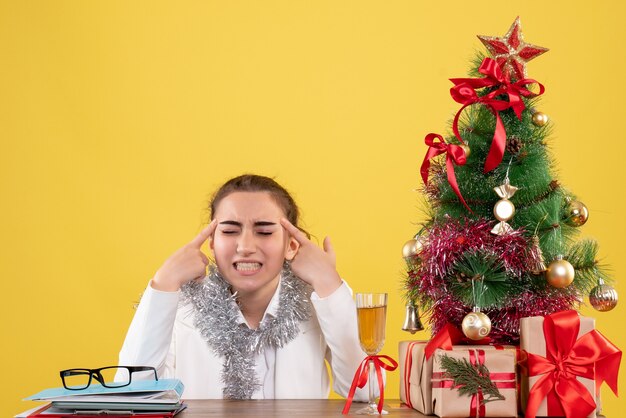 Vista frontal doctora sentada detrás de su mesa sobre fondo amarillo con árbol de navidad y cajas de regalo