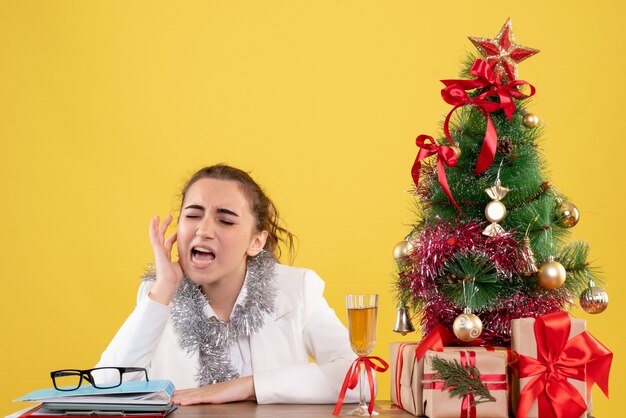 Vista frontal doctora sentada detrás de su mesa sobre fondo amarillo con árbol de navidad y cajas de regalo