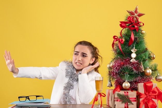 Vista frontal doctora sentada detrás de su mesa sobre fondo amarillo con árbol de navidad y cajas de regalo