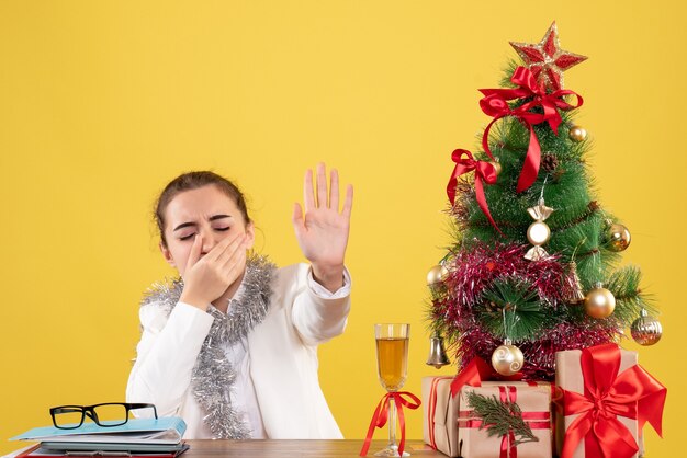 Vista frontal doctora sentada detrás de su mesa sobre fondo amarillo con árbol de navidad y cajas de regalo