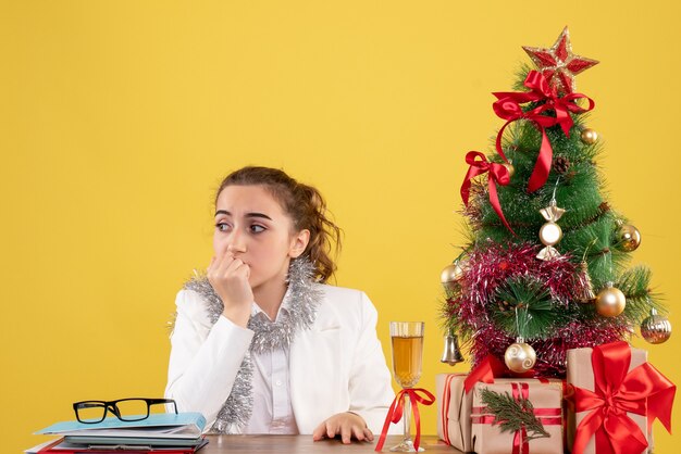 Vista frontal doctora sentada detrás de su mesa sobre fondo amarillo con árbol de navidad y cajas de regalo