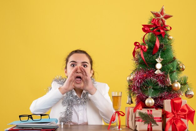 Vista frontal doctora sentada detrás de su mesa sobre fondo amarillo con árbol de navidad y cajas de regalo