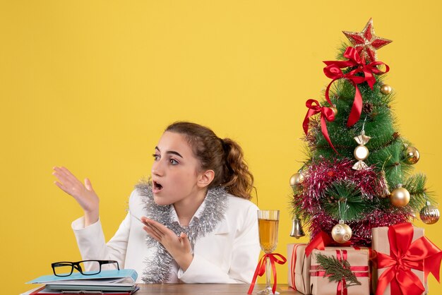 Vista frontal doctora sentada detrás de su mesa sobre fondo amarillo con árbol de navidad y cajas de regalo