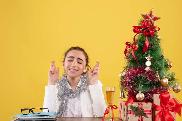 Vista frontal doctora sentada detrás de su mesa sobre fondo amarillo con árbol de navidad y cajas de regalo