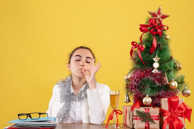Vista frontal doctora sentada detrás de su mesa sobre fondo amarillo con árbol de navidad y cajas de regalo