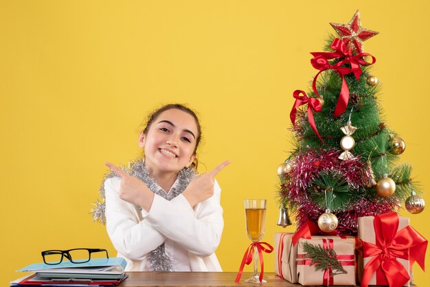 Vista frontal doctora sentada detrás de su mesa sonriendo sobre fondo amarillo con árbol de navidad y cajas de regalo