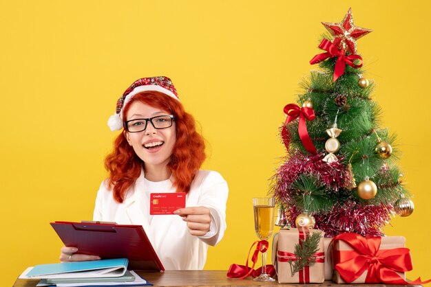 Vista frontal doctora sentada detrás de su mesa y sosteniendo una tarjeta bancaria sobre fondo amarillo con árbol de navidad y cajas de regalo