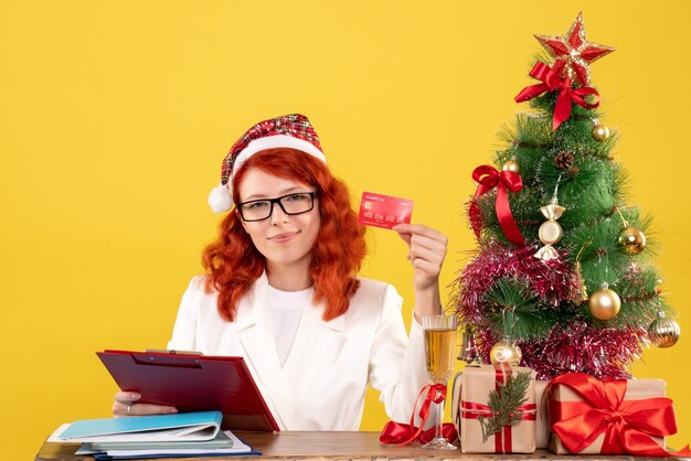 Vista frontal doctora sentada detrás de su mesa y sosteniendo una tarjeta bancaria sobre fondo amarillo con árbol de navidad y cajas de regalo