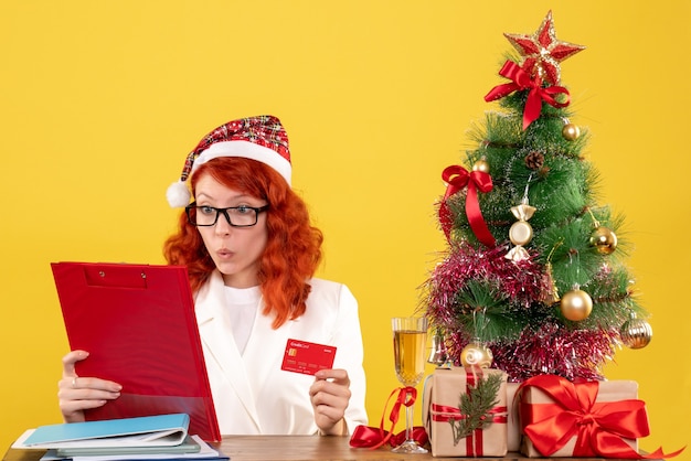 Vista frontal doctora sentada detrás de su mesa y sosteniendo una tarjeta bancaria sobre fondo amarillo con árbol de navidad y cajas de regalo