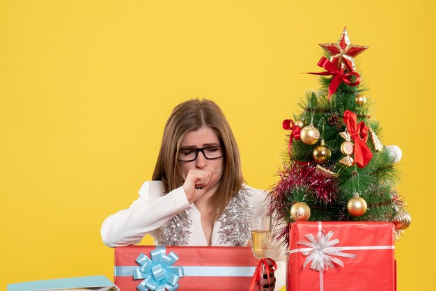 Vista frontal doctora sentada frente a la mesa con regalos y árbol en el escritorio amarillo