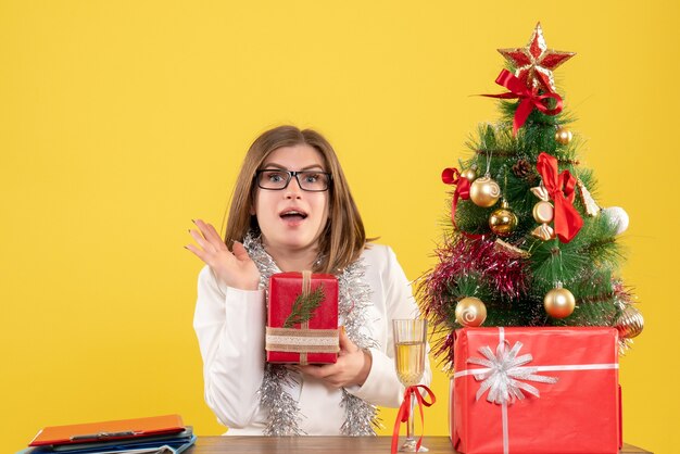 Vista frontal doctora sentada frente a la mesa con regalos y árbol sobre fondo amarillo