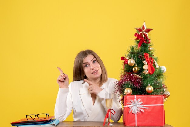 Vista frontal doctora sentada frente a la mesa con regalos de Navidad y árbol sobre fondo amarillo