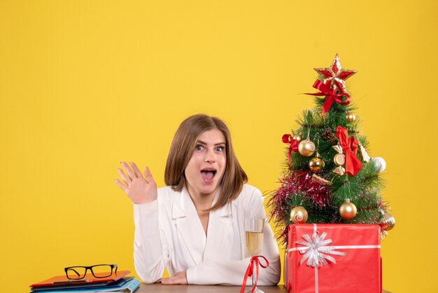 Vista frontal doctora sentada frente a la mesa con regalos de Navidad y árbol sobre fondo amarillo
