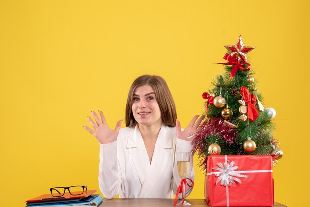Vista frontal doctora sentada frente a la mesa con regalos de Navidad y árbol sobre fondo amarillo