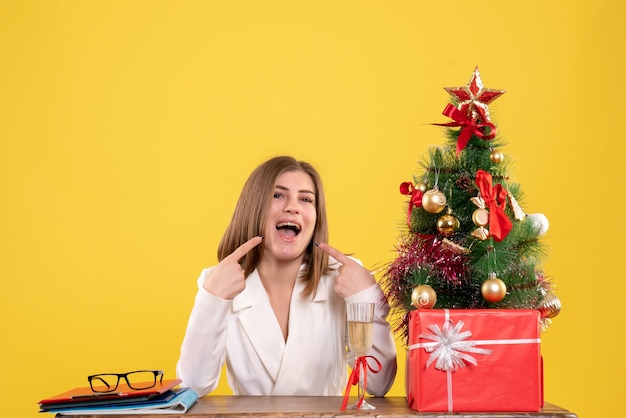 Vista frontal doctora sentada frente a la mesa con regalos de Navidad y árbol sobre fondo amarillo