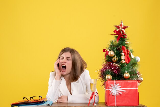 Vista frontal doctora sentada frente a su mesa bostezando sobre fondo amarillo con árbol de navidad y cajas de regalo