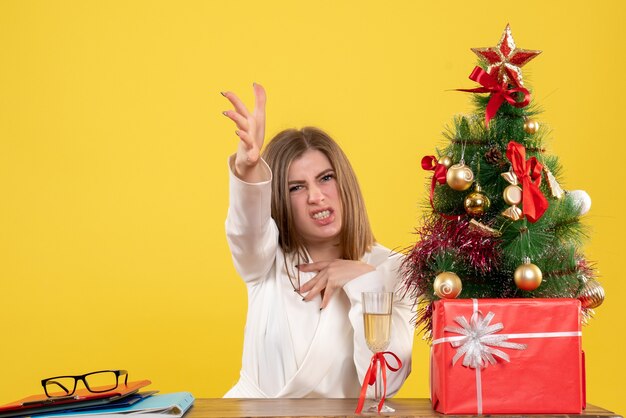 Vista frontal doctora sentada frente a su mesa disgustada sobre fondo amarillo con árbol de navidad y cajas de regalo