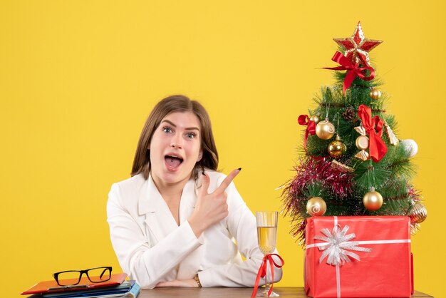 Vista frontal doctora sentada frente a su mesa en el escritorio amarillo con árbol de navidad y cajas de regalo