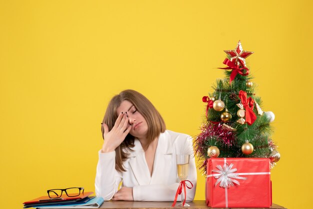 Vista frontal doctora sentada frente a su mesa en el escritorio amarillo con árbol de navidad y cajas de regalo