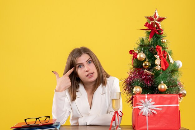 Vista frontal doctora sentada frente a su mesa en el escritorio amarillo con árbol de navidad y cajas de regalo