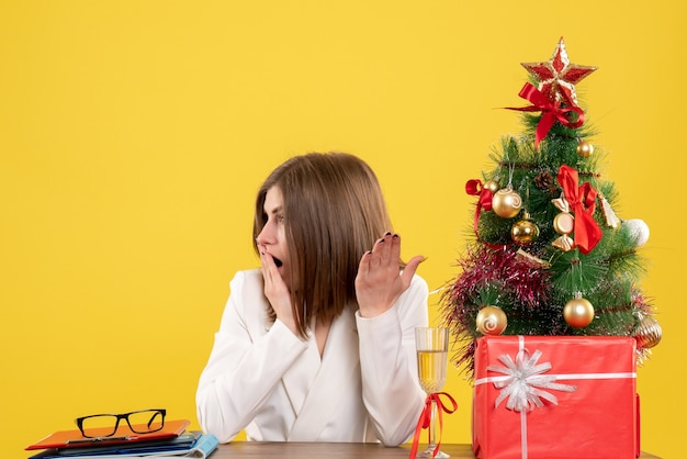 Vista frontal doctora sentada frente a su mesa sobre un fondo amarillo con árbol de navidad y cajas de regalo