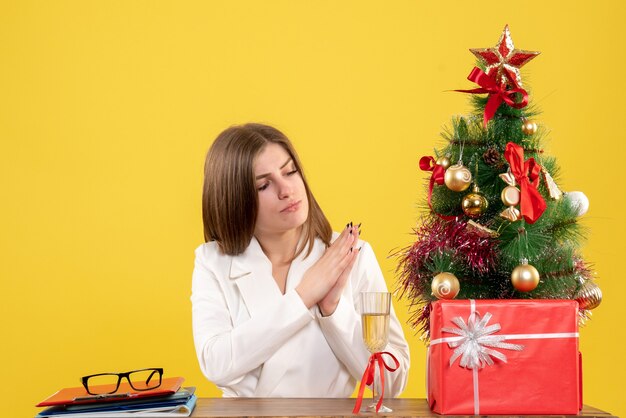 Vista frontal doctora sentada frente a su mesa sobre un fondo amarillo con árbol de navidad y cajas de regalo