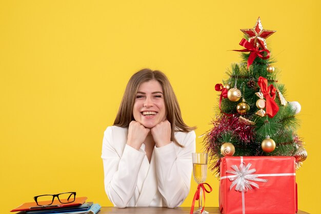 Vista frontal doctora sentada frente a su mesa sobre un fondo amarillo con árbol de navidad y cajas de regalo