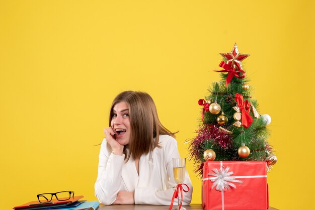 Vista frontal doctora sentada frente a su mesa sobre un fondo amarillo con árbol de navidad y cajas de regalo