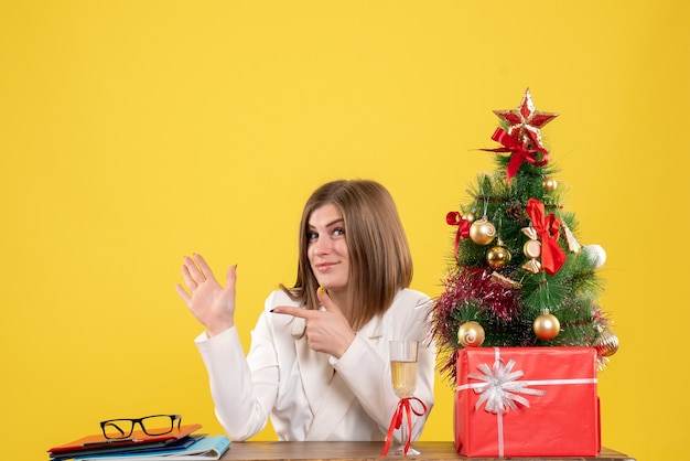 Vista frontal doctora sentada frente a su mesa sobre un fondo amarillo con árbol de navidad y cajas de regalo