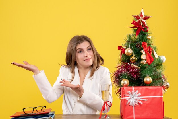 Vista frontal doctora sentada frente a su mesa sobre un fondo amarillo con árbol de navidad y cajas de regalo