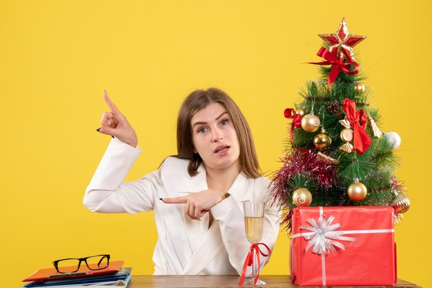 Vista frontal doctora sentada frente a su mesa sobre un fondo amarillo con árbol de navidad y cajas de regalo