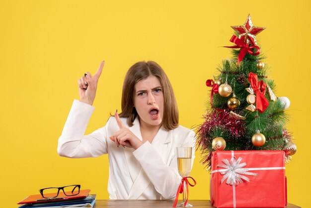 Vista frontal doctora sentada frente a su mesa sobre un fondo amarillo con árbol de navidad y cajas de regalo