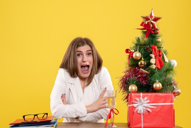 Vista frontal doctora sentada frente a su mesa sobre un fondo amarillo con árbol de navidad y cajas de regalo