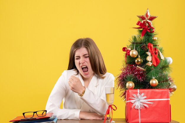 Vista frontal doctora sentada frente a su mesa sobre un fondo amarillo con árbol de navidad y cajas de regalo