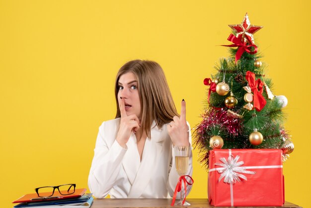 Vista frontal doctora sentada frente a su mesa sobre un fondo amarillo con árbol de navidad y cajas de regalo