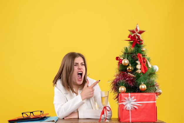 Vista frontal doctora sentada frente a su mesa sobre un fondo amarillo con árbol de navidad y cajas de regalo