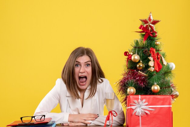 Vista frontal doctora sentada frente a su mesa sobre un fondo amarillo con árbol de navidad y cajas de regalo