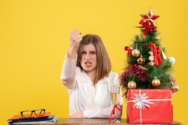 Vista frontal doctora sentada frente a su mesa sobre un fondo amarillo con árbol de navidad y cajas de regalo