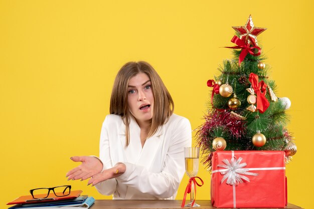 Vista frontal doctora sentada frente a su mesa sobre un fondo amarillo con árbol de navidad y cajas de regalo