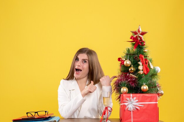 Vista frontal doctora sentada frente a su mesa sobre un fondo amarillo con árbol de navidad y cajas de regalo