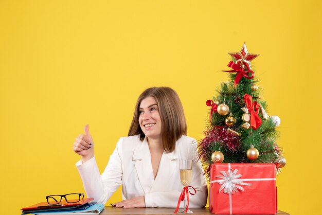 Vista frontal doctora sentada frente a su mesa sobre un fondo amarillo con árbol de navidad y cajas de regalo