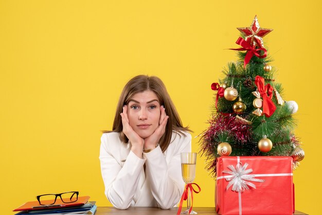 Vista frontal doctora sentada frente a su mesa sobre un fondo amarillo con árbol de navidad y cajas de regalo