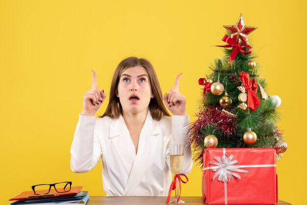 Vista frontal doctora sentada frente a su mesa sobre un fondo amarillo con árbol de navidad y cajas de regalo