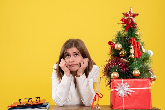 Vista frontal doctora sentada frente a su mesa sobre un fondo amarillo con árbol de navidad y cajas de regalo