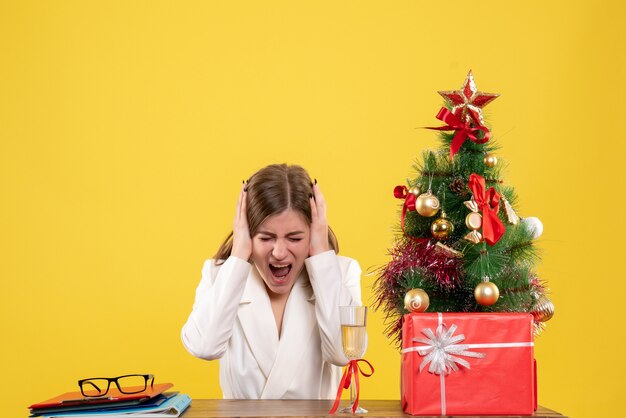 Vista frontal doctora sentada frente a su mesa sobre un fondo amarillo con árbol de navidad y cajas de regalo