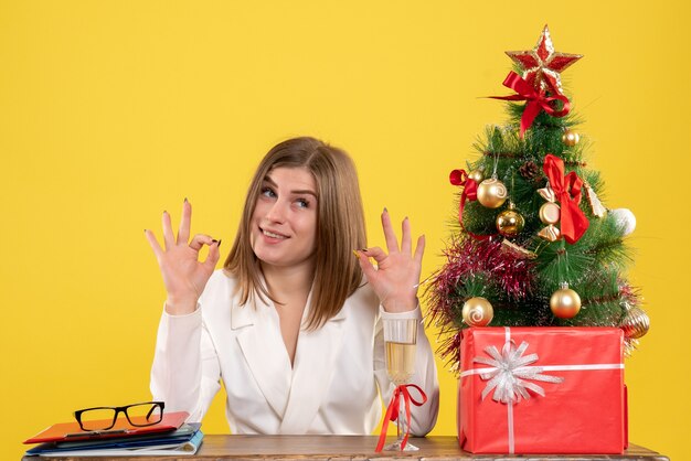 Vista frontal doctora sentada frente a su mesa sobre un fondo amarillo con árbol de navidad y cajas de regalo